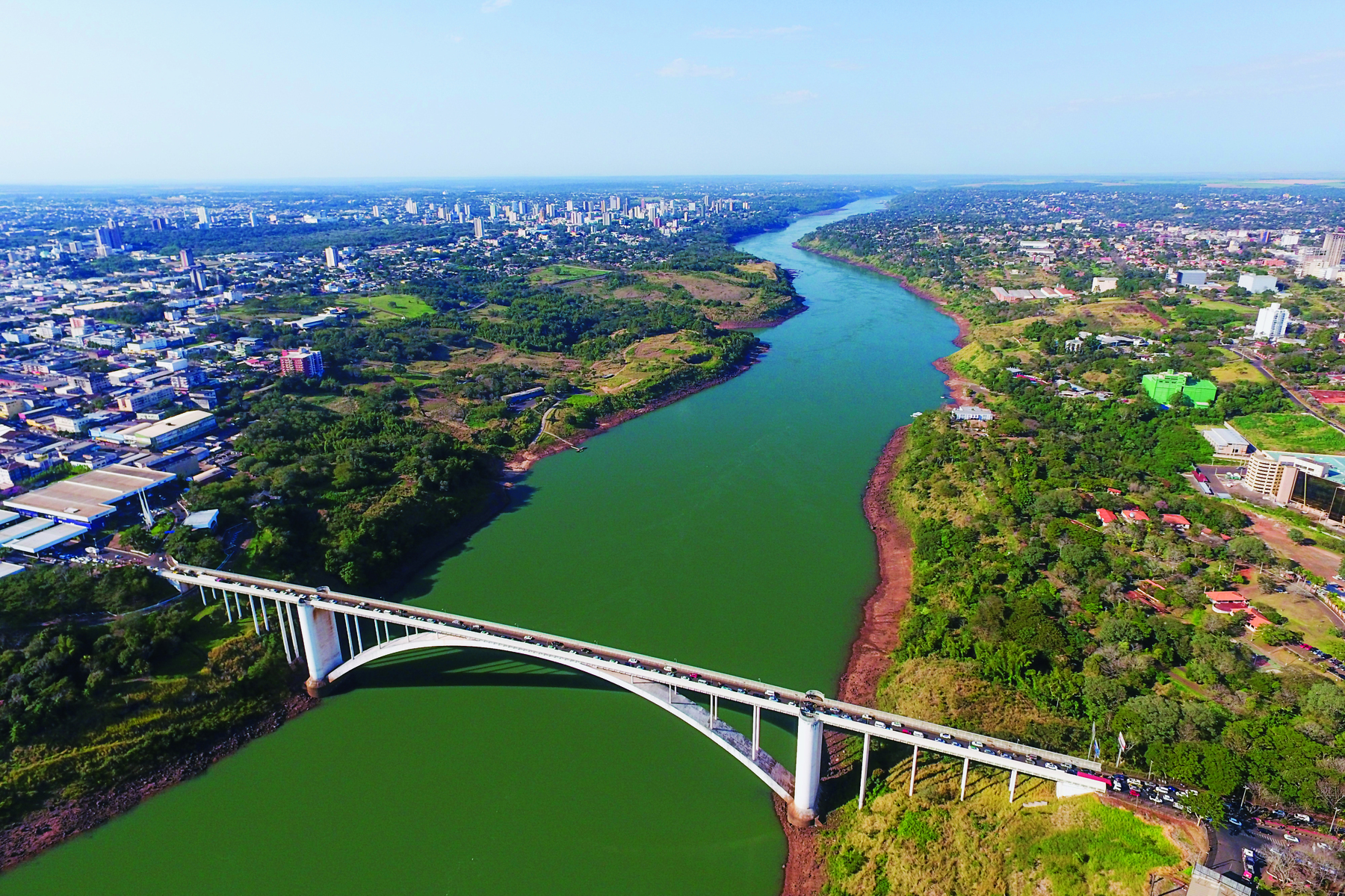 Fotografia. Vista de uma ponte com a base em fórma de arco sobre um rio extenso e largo. As margens têm vegetação. Após essa vegetação há diversas construções em  ambas as margens.