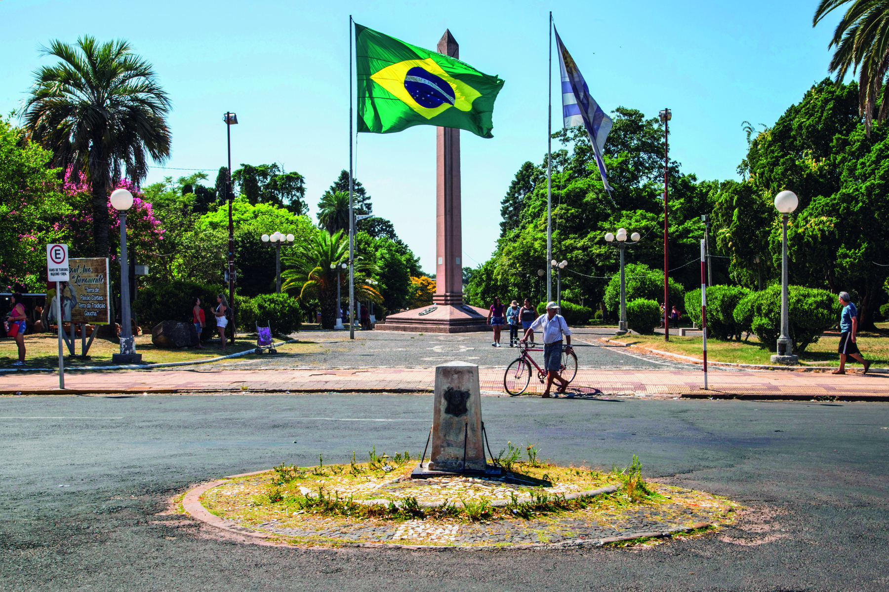 Fotografia. Vista de uma praça com várias árvores e  um grande monumento vertical de concreto no centro. Em frente há dois mastros, um ao lado do outro. No mastro à esquerda está a  bandeira do Brasil; no mastro à direita, a bandeira do Uruguai. Em frente, uma rua asfaltada com uma pessoa de bicicleta.