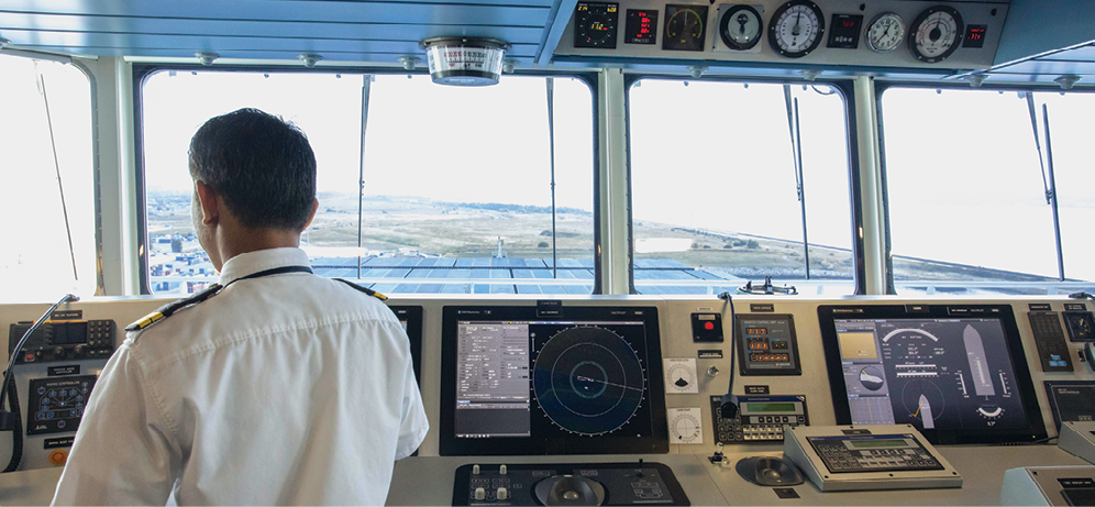 Fotografia. Homem de uniforme no interior da cabine de comando de um navio. Ele está de costas, de frente para um grande painel com diversas telas e botões. Acima do painel estão diversas janelas de vidro de onde se pode avistar uma área litorânea.