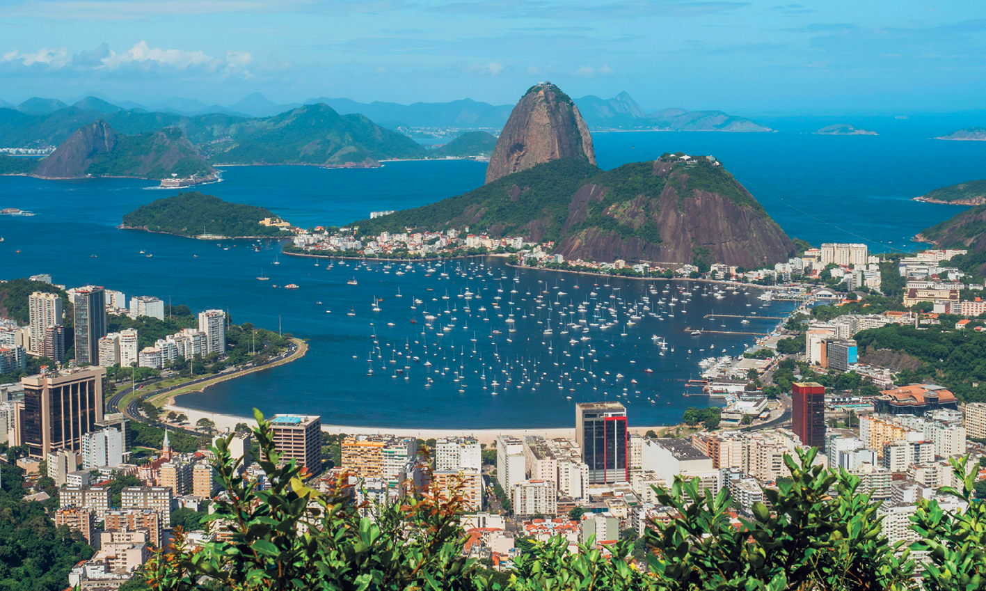 Fotografia. No centro da foto, vista dos morros Pão de Açúcar e da Urca: dois morros próximos, sendo o do Pão de Açúcar mais alto e pontiagudo e, o da Urca, menor e mais largo. Aos pés dos morros há muitas construções. Ao redor dos morros e ao fundo, o mar e outros morros. À frente, faixa de areia da praia com muitos prédios ao longo de sua extensão e alguma vegetação.