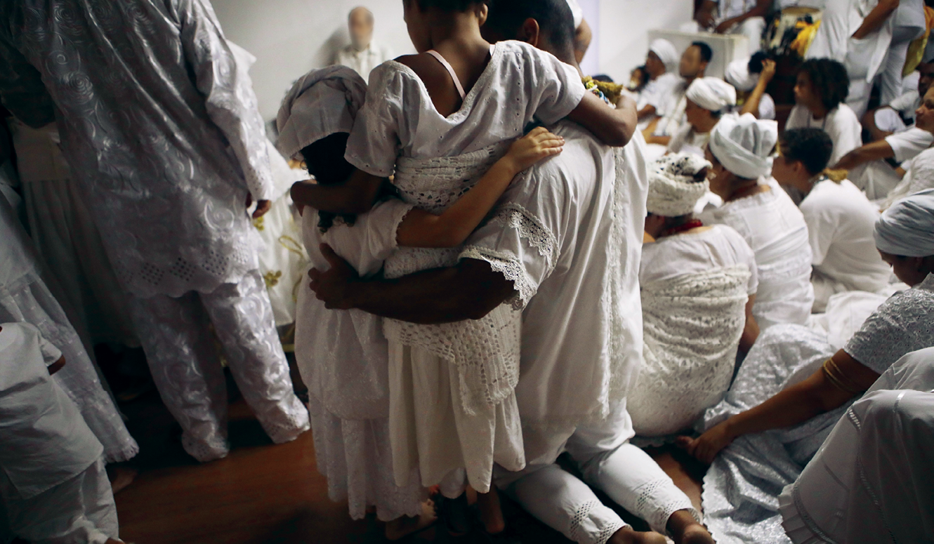 Fotografia. Grupo de pessoas vestidas com roupas brancas, longas e com babados. Algumas pessoas usam pano branco amarrado na cabeça e nas costas. Elas estão participando de um ritual religioso.