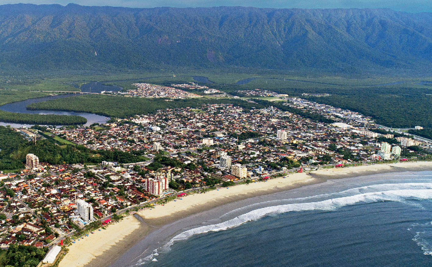 Fotografia. Vista aérea de uma cidade à beira mar. No primeiro plano, o mare uma faixa de areia. Na sequência, uma área urbana com muitas construções. Ao fundo, um rio, muita vegetação e grandes montanhas cobertas de vegetação.