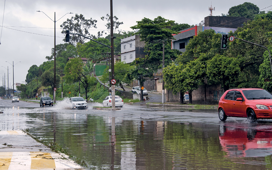 Fotografia. Vista de uma rua alagada. No primeiro plano, alguns carros atravessando a área alagada. No segundo plano, algumas árvores plantadas ao longo da calçada e diversas construções.