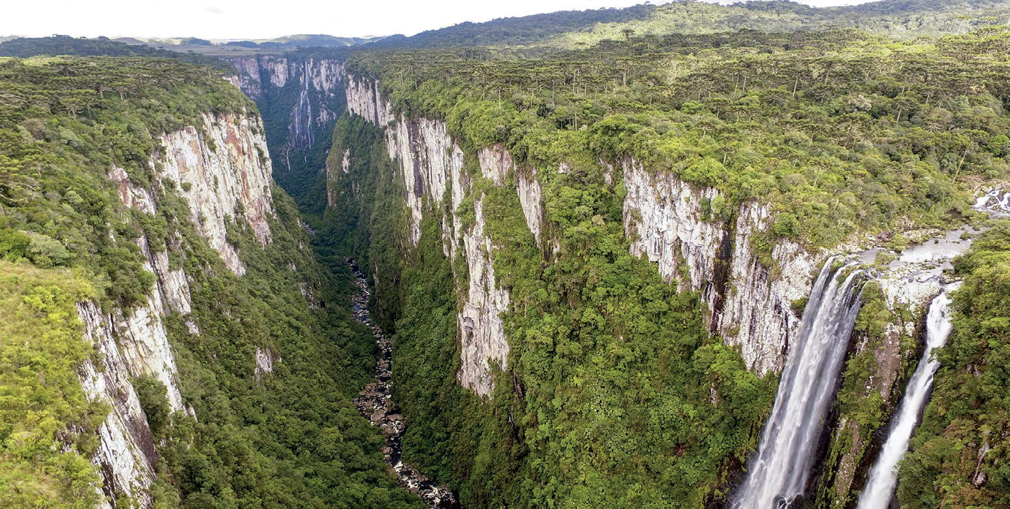 Fotografia. Vista do alto mostrando uma grande fenda aberta entre paredões rochosos, formando um extenso vale. À direita, uma cachoeira despejando água no rio, que corre no fundo do vale. Ao longo do vale e acima dos paredões, há vasta vegetação.