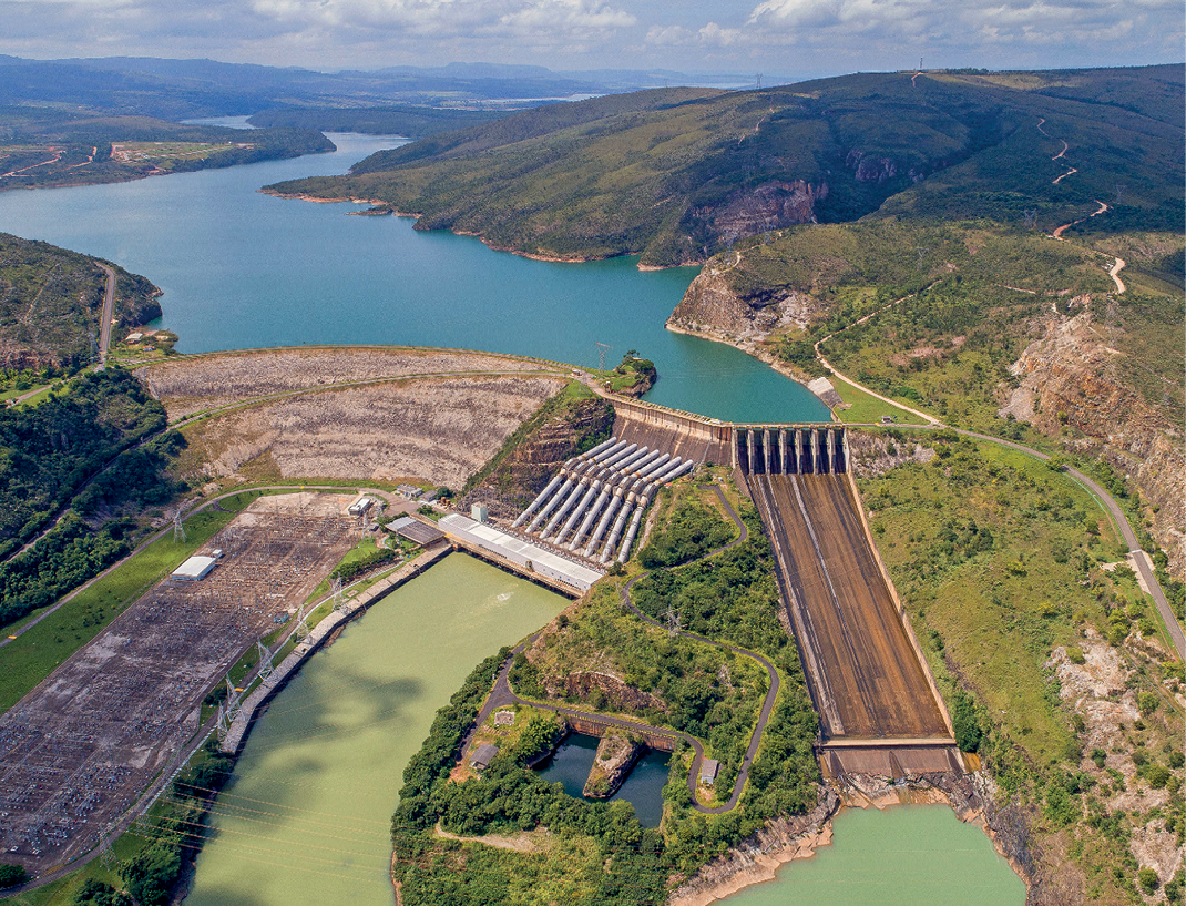 Fotografia. Em primeiro plano, há uma usina hidrelétrica, na qual é possível observar a barragem, outras estruturas de concreto e parte de um curso de água represado. Em segundo plano, em uma área mais elevada e ligado à usina, extenso rio margeado por um morro que ocupa a maior parte do lado direito da fotografia.