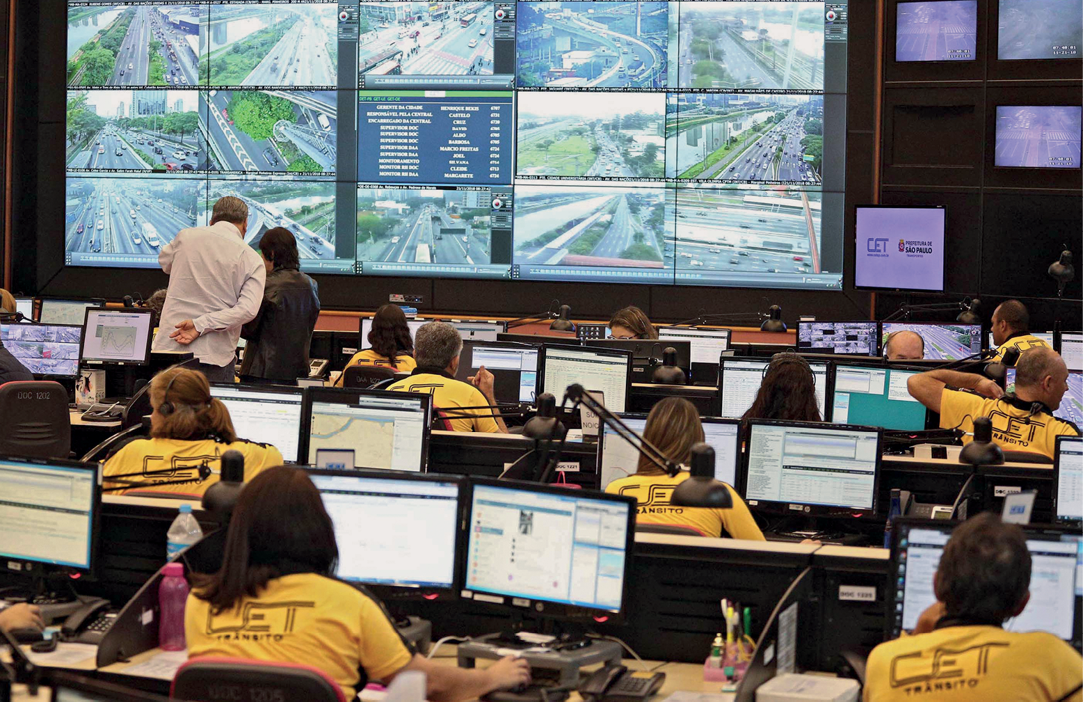 Fotografia. Vista de uma sala ampla com várias telas em uma parede transmitindo imagens das vias de circulação monitoradas. À frente das telas estão diversas pessoas sentadas em mesas com computadores. Elas usam uniformes e observam as telas.