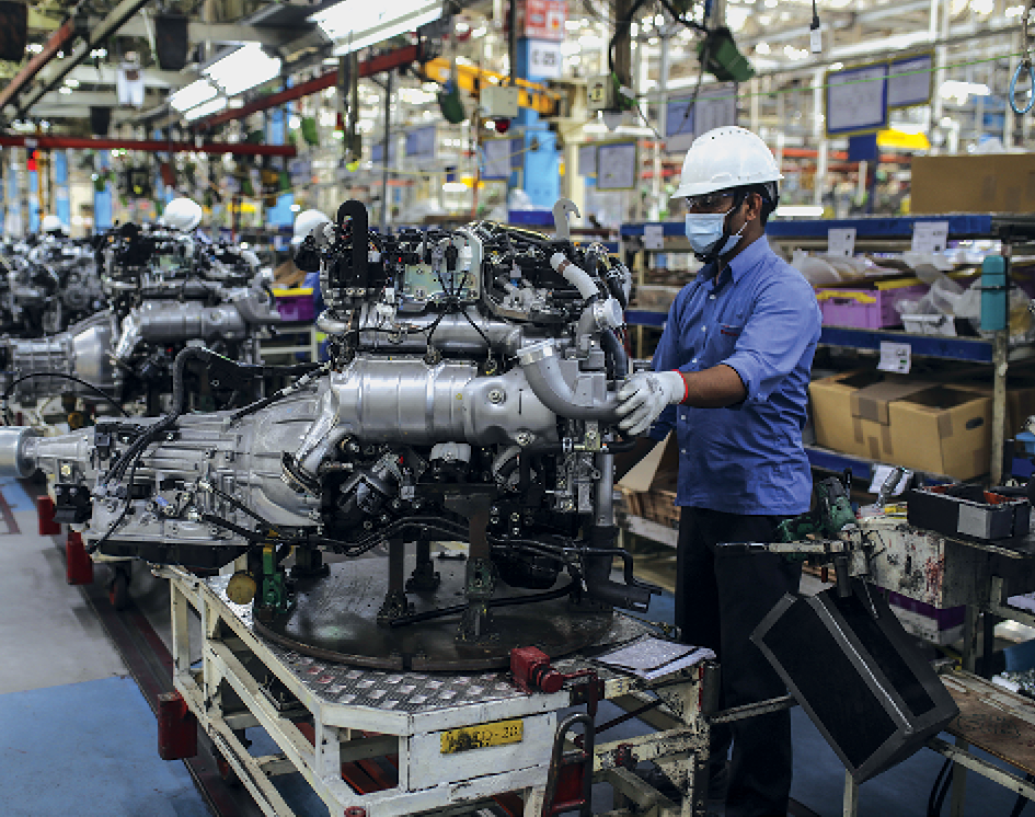 Fotografia. Vista do interior de uma fábrica de motores. Destaque para uma pessoa de uniforme e capacete manuseando ferramentas e trabalhando na montagem de um motor. Há muitos fios e peças expostas.