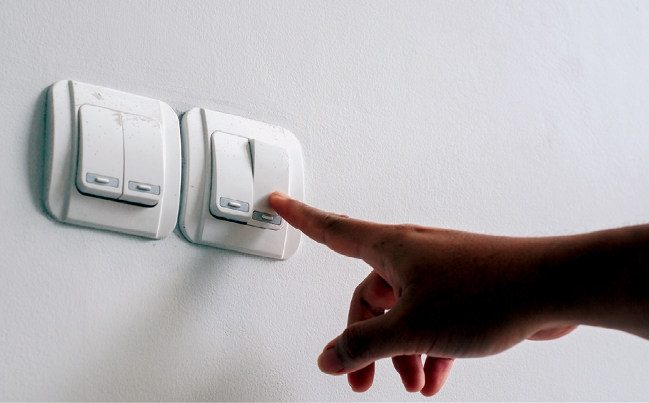 Fotografia. Uma pessoa apertando o botão que um interruptor na parede. Há quatro botões para ascender e apagar a luz.