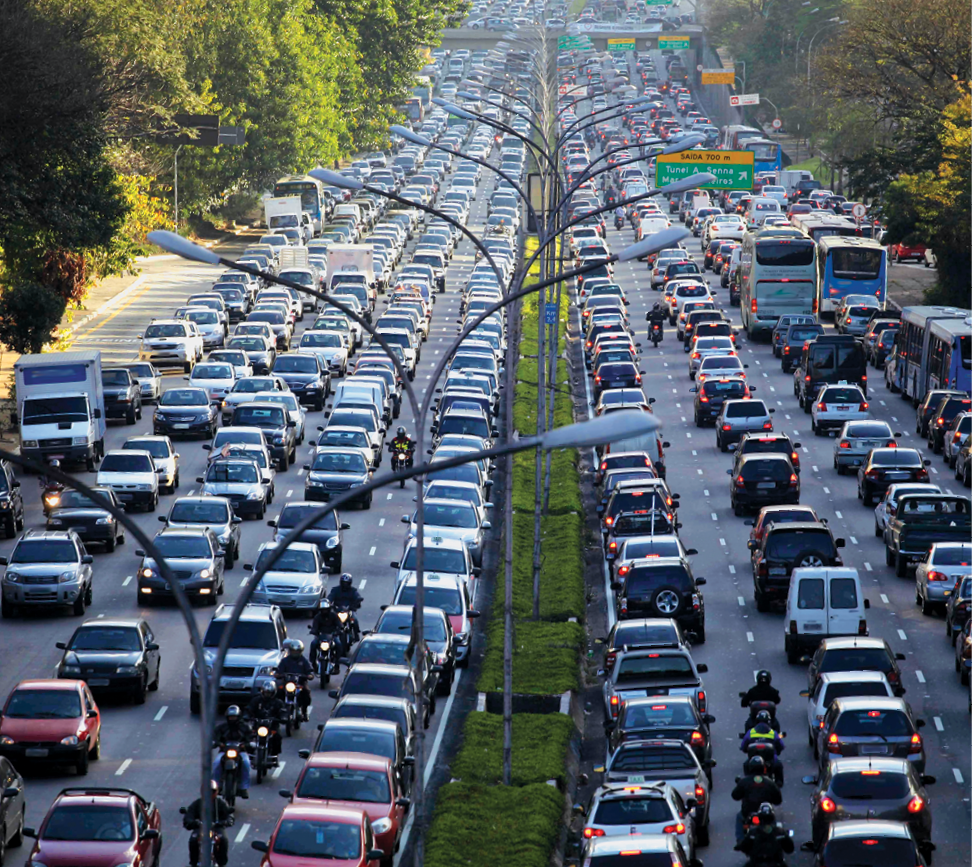 Fotografia. Vista de uma avenida com cinco faixas de rolamento em cada pista. Há muitos veículos em ambas as direções, em um situação de congestionamento.