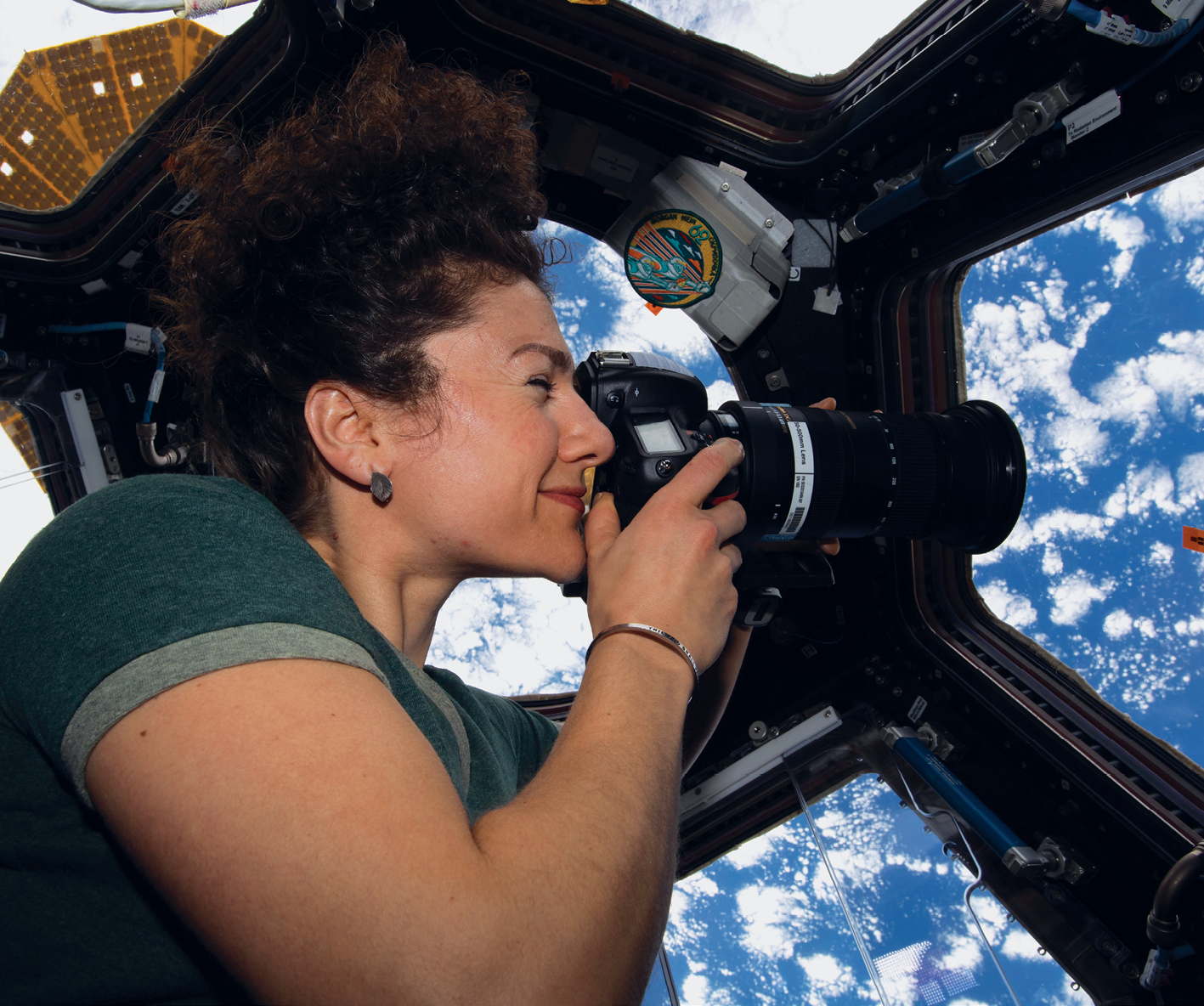 Fotografia. Uma mulher dentro de uma estação espacial, com as laterais feitas de diversas janelas de vidro. Ela é branca, tem cabelo encaracolado preso no alto da cabeça, segura uma máquina fotográfica próxima aos olhos e aponta a máquina para uma das janelas. Do lado de fora da estação há  diversas nuvens e céu azul.