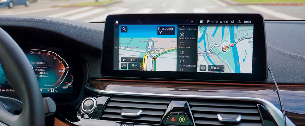 Fotografia. Painel de um carro com destaque para a tela de um equipamento de GPS que mostra um trajeto em um mapa colorido.
