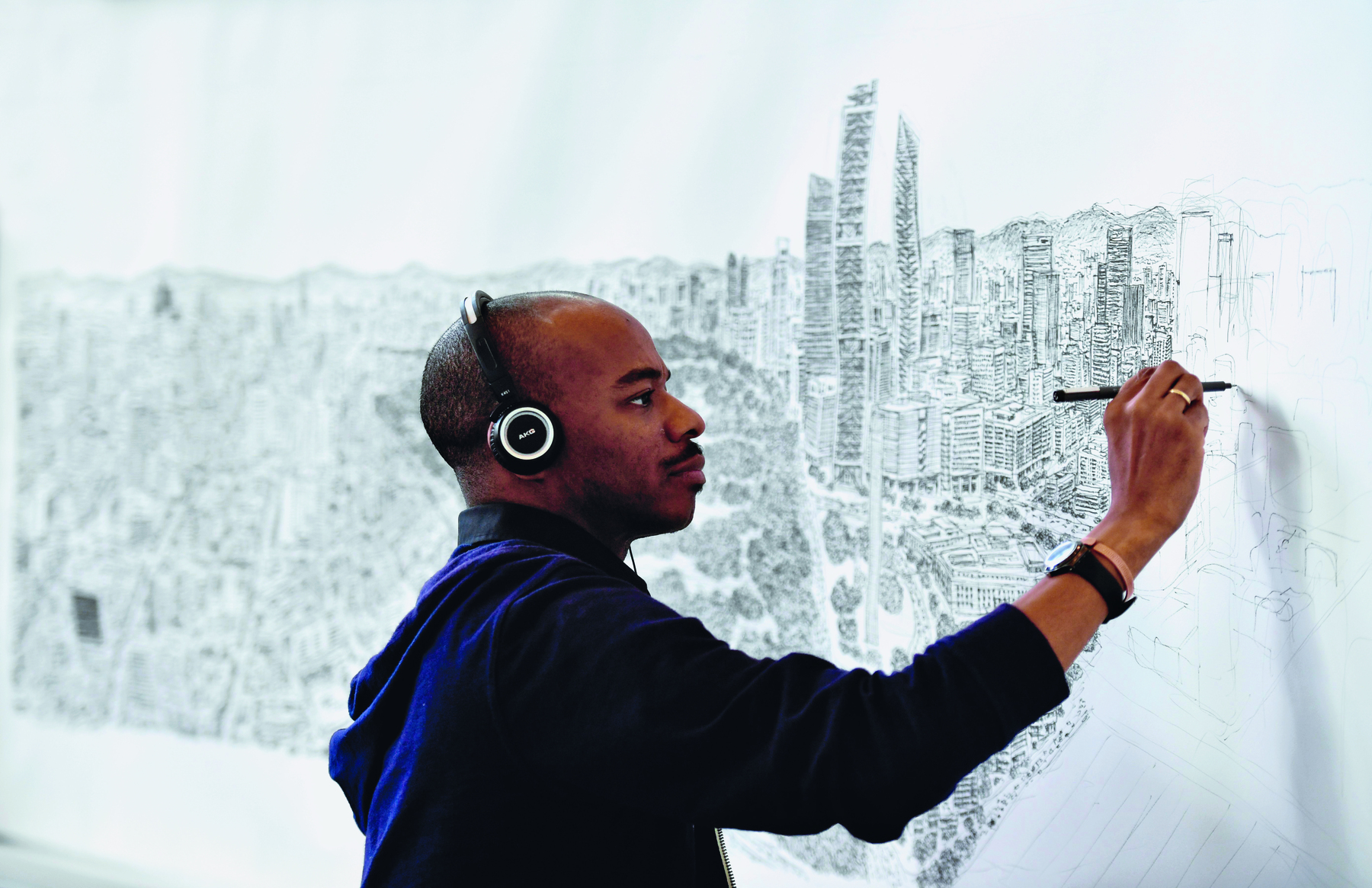 Fotografia. Um homem  visto de perfil da cintura para cima. Ele é negro, usa camisa azul, fone nos ouvidos e desenha as construções de uma cidade em um painel.