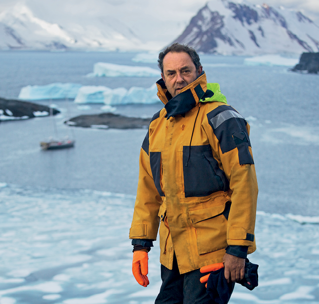 Fotografia de um homem branco, cabelos curtos, de jaqueta longa, luvas e calça. Ao redor dele, uma faixa de água com placas de gelo. Ao fundo, montanhas cobertas de gelo.