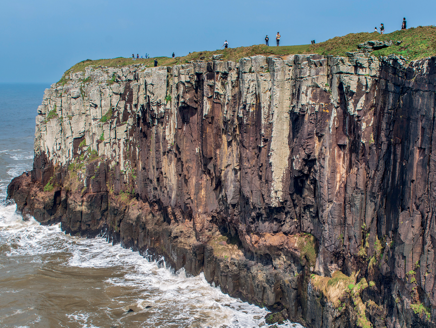 Fotografia. Vista de uma grande formação rochosa de frente para o oceano, com a feição de um paredão à beira mar. No topo, acima do paredão, há diversas pessoas observando.