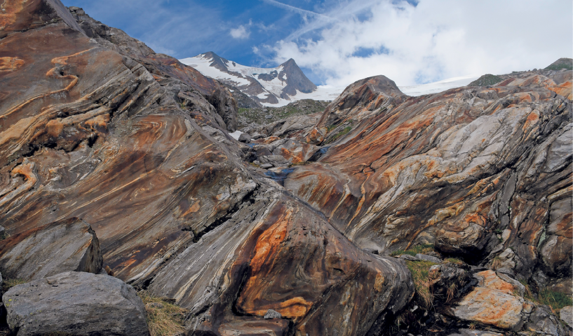 Fotografia. Vista de uma formação rochosa em uma área montanhosa. O desenho das rochas apresenta formas com ondulações, que são encurvamentos das dobras.