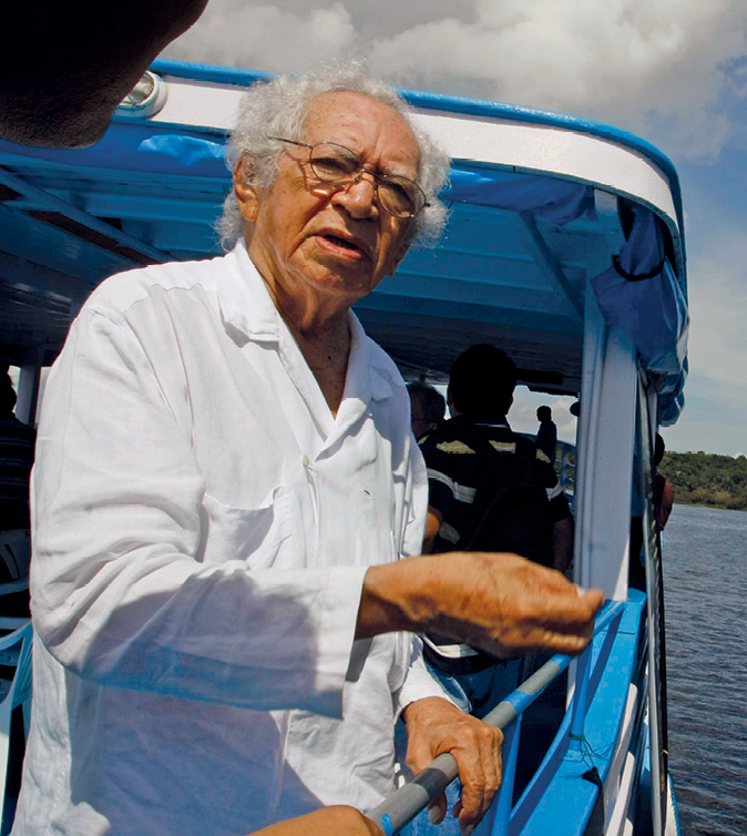 Fotografia de um idoso, de cabelos brancos e usando óculos e camisa branca. Ele está dentro de um barco azul e branco que está em um rio.