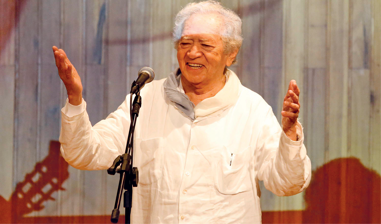 Fotografia de um idoso, de cabelos brancos e usando camisa branca. Ele está na frente de um microfone, sorrindo, e com os braços abertos.