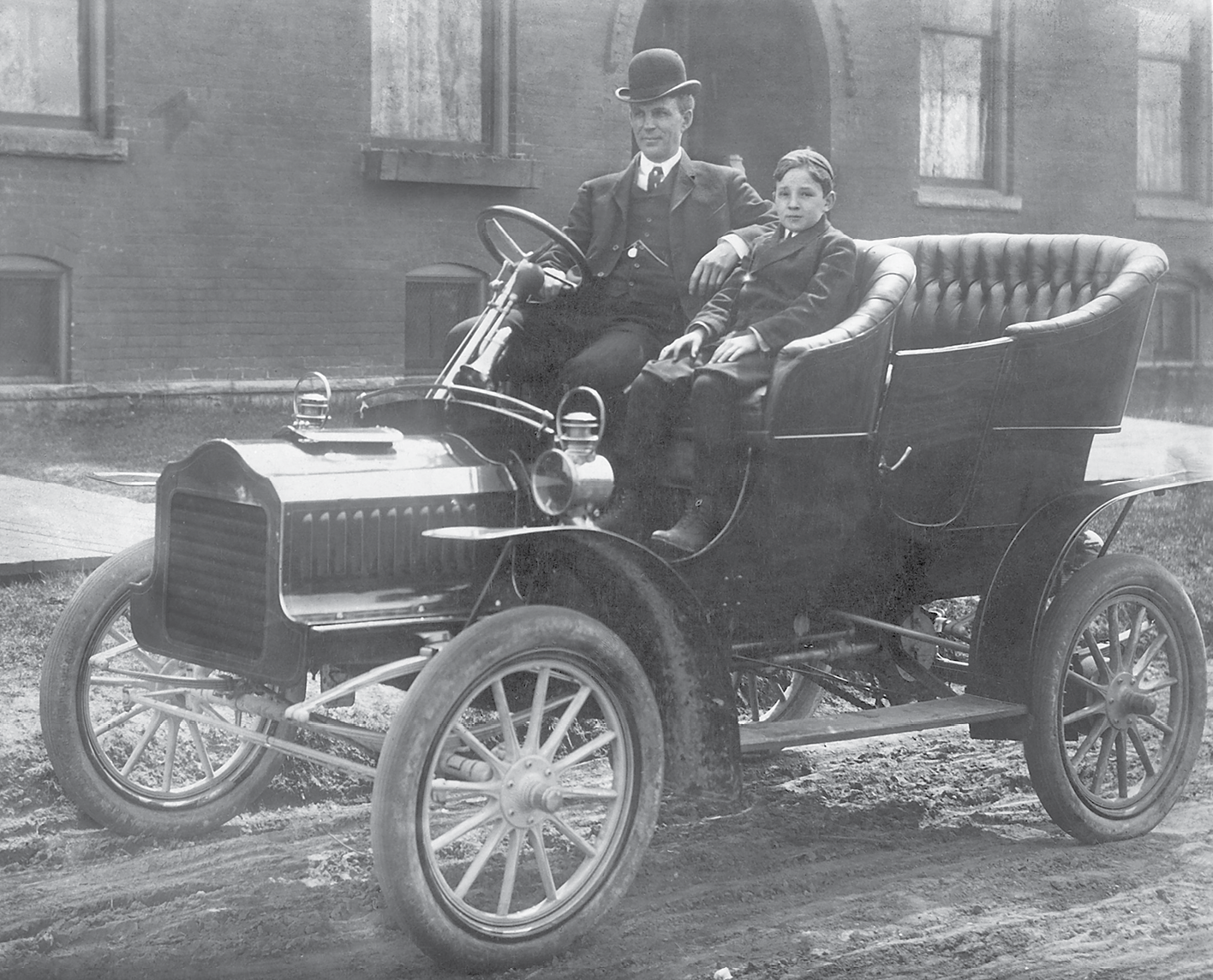 Fotografia em preto e branco. Um homem de terno, gravata e chapéu e uma criança pequena. Eles posam para foto dentro de um carro. O carro tem rodas finas, não possui teto e o volante tem a base comprida.