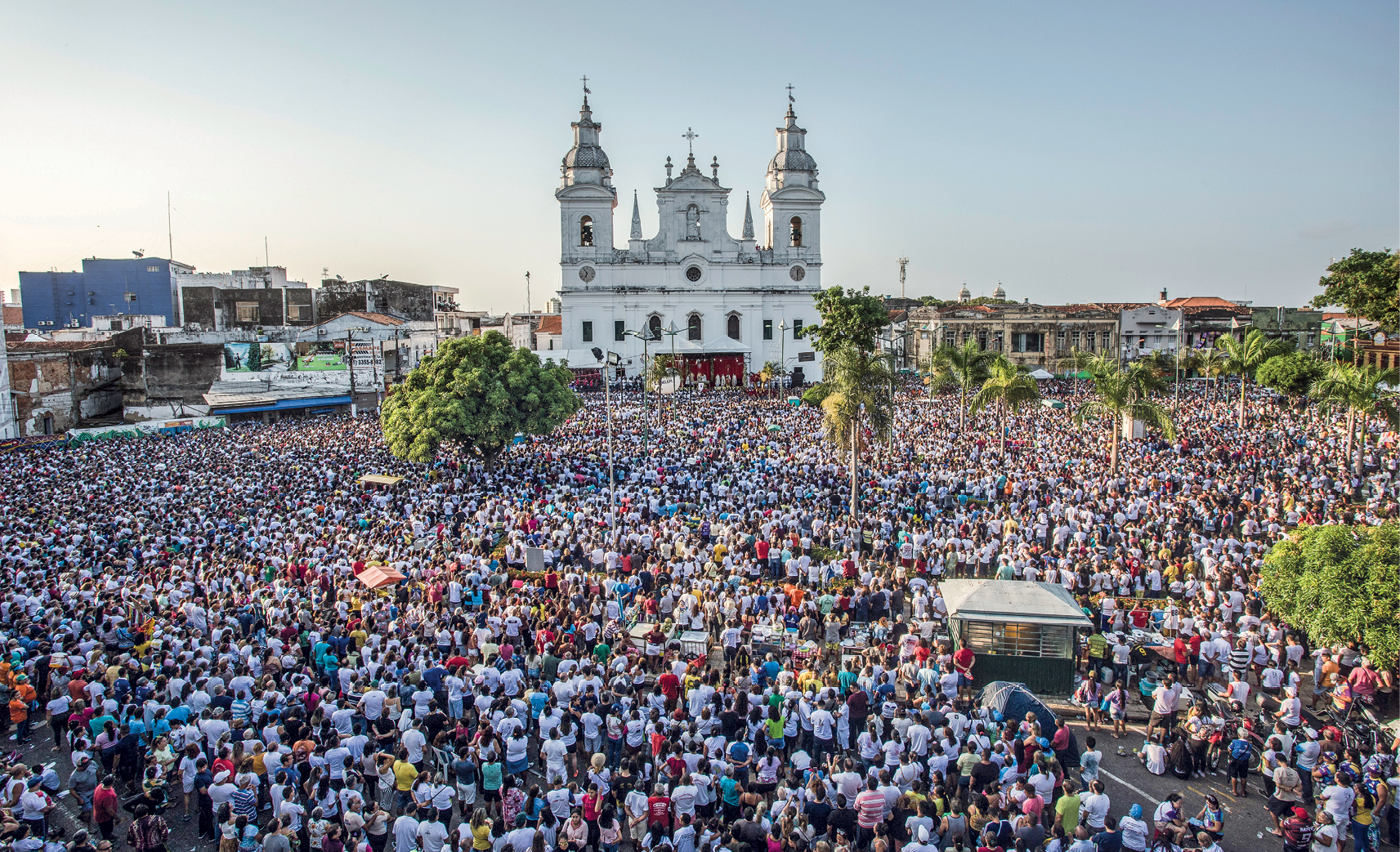 Fotografia. Vista de uma praça ampla com uma multidão de pessoas. Elas estão olhando em direção a um palco que está na frente de uma grande igreja com duas torres e cruzes no topo.