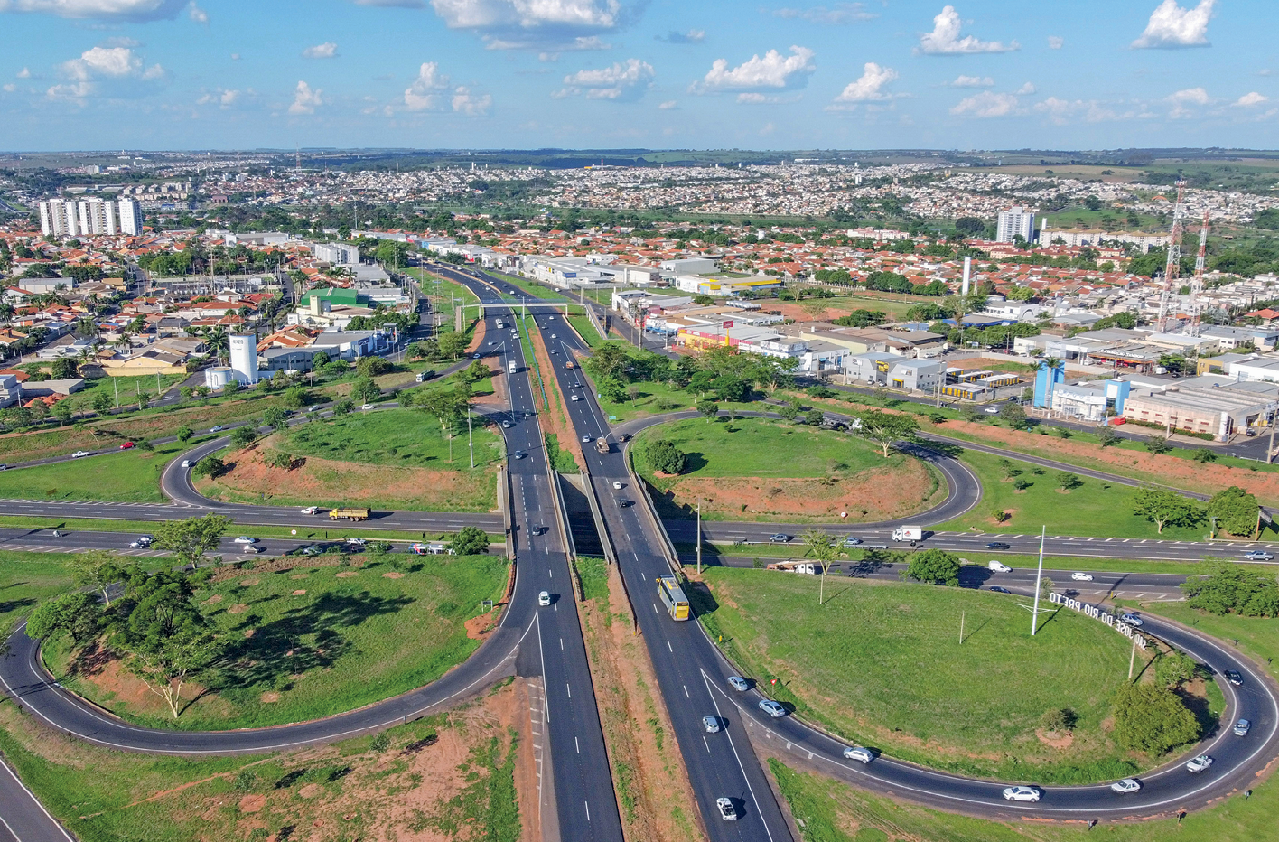 Fotografia. No centro da imagem, um viaduto com duas pistas em cada sentido. Sob o viaduto, há uma rodovia formada por duas pistas, em cada sentido, separadas por um canteiro central. O viaduto e a rodovia estão conectados por meio de quatro rotatórias. Na parte superior, à direita e à esquerda, há construções predominantemente baixas, alguns prédios, galpões, áreas asfaltadas e árvores.