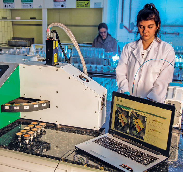 Fotografia. Mulher de jaleco branco próxima a um equipamento branco e a um notebook aberto. Ao fundo, atrás de uma bancada com diversos objetos, há outra mulher.