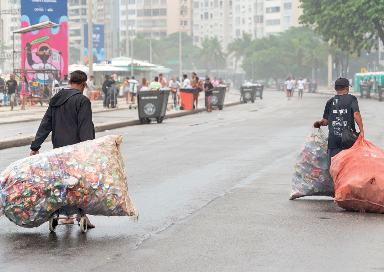 Fotografia. Em primeiro plano, duas pessoas aparecem de costas carregando grandes sacos com latas. Em segundo plano, diversas pessoas caminhando na calçada, árvores e prédios.
