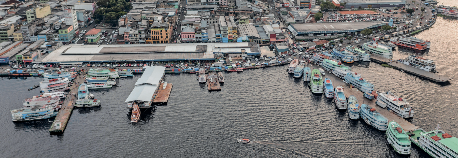 Fotografia. Vista de um porto com muitas embarcações ancoradas. Nas margens estão diversos galpões e uma rua com muitas pessoas e carros. Ao fundo, construções baixas e alguns prédios.