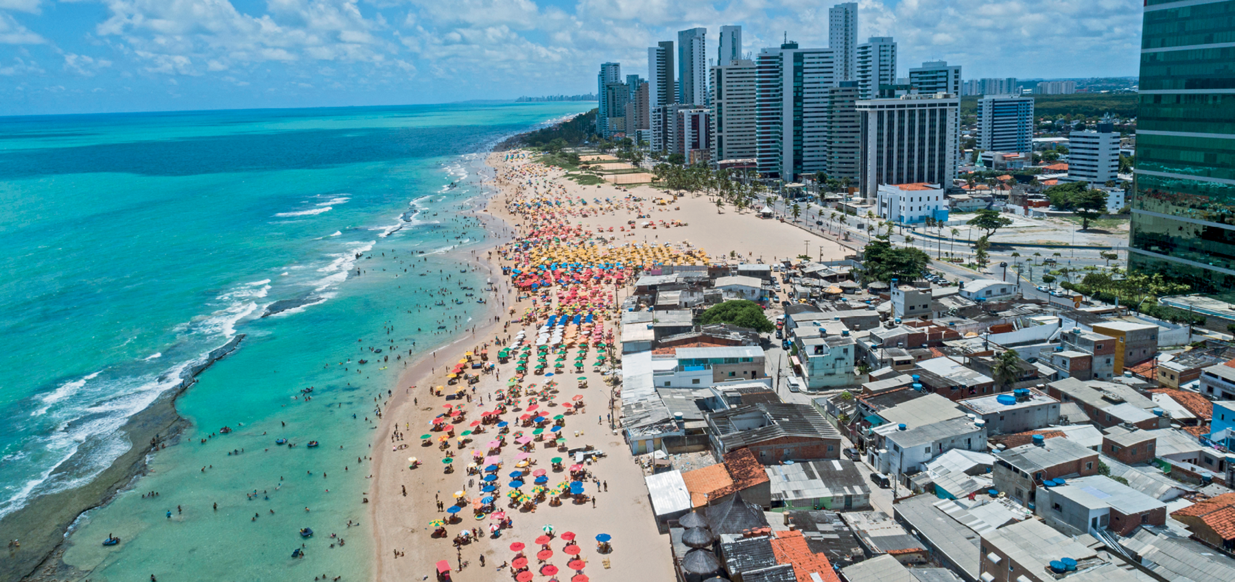 Fotografia. Vista da área litorânea de uma cidade em dia ensolarado. No centro, faixa de areia com diversas pessoas e guarda-sóis. À esquerda, o mar refletindo a cor azul, com muitos banhistas na água; à direita, uma avenida de frente para a praia e construções de diferentes tipos, desde casas à edifícios variados.