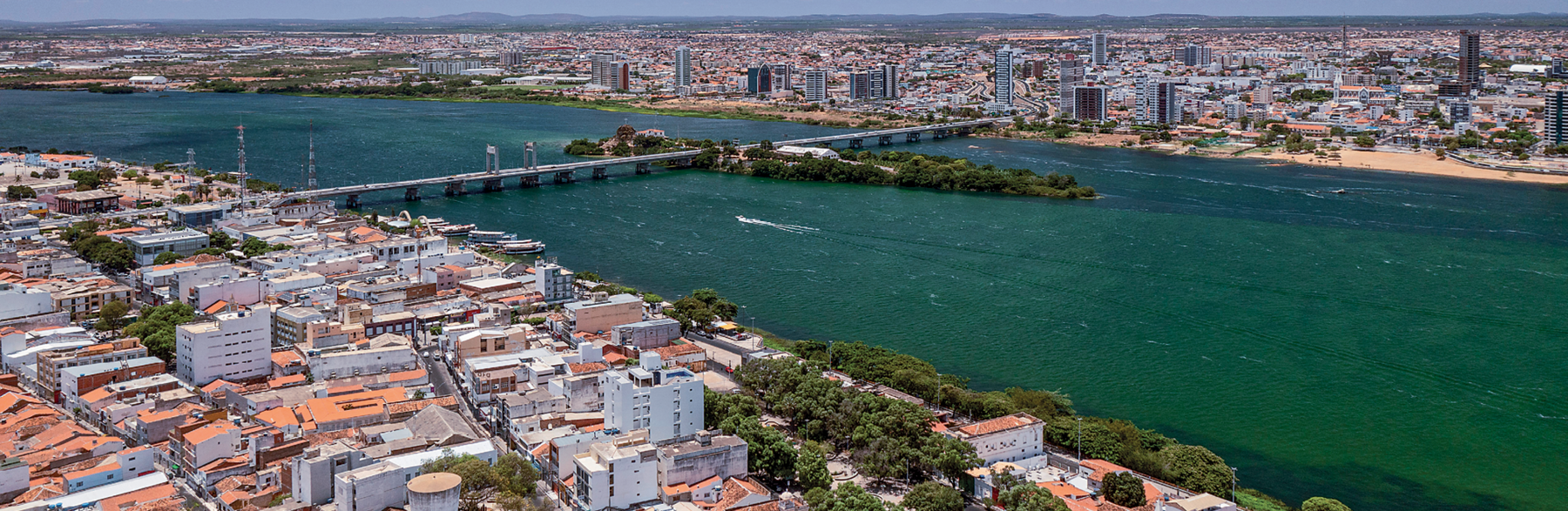 Fotografia. Vista de um rio caudaloso, de extensão larga, no centro da imagem, atravessando da esquerda para a direita. Na parte de cima, ao fundo, junto a uma das margens do rio, aparece uma cidade. Na parte de baixo da fotografia, junto a outra margem do rio, aparece mais uma cidade. Existe uma ponte que atravessa o rio, conectando as duas cidades.