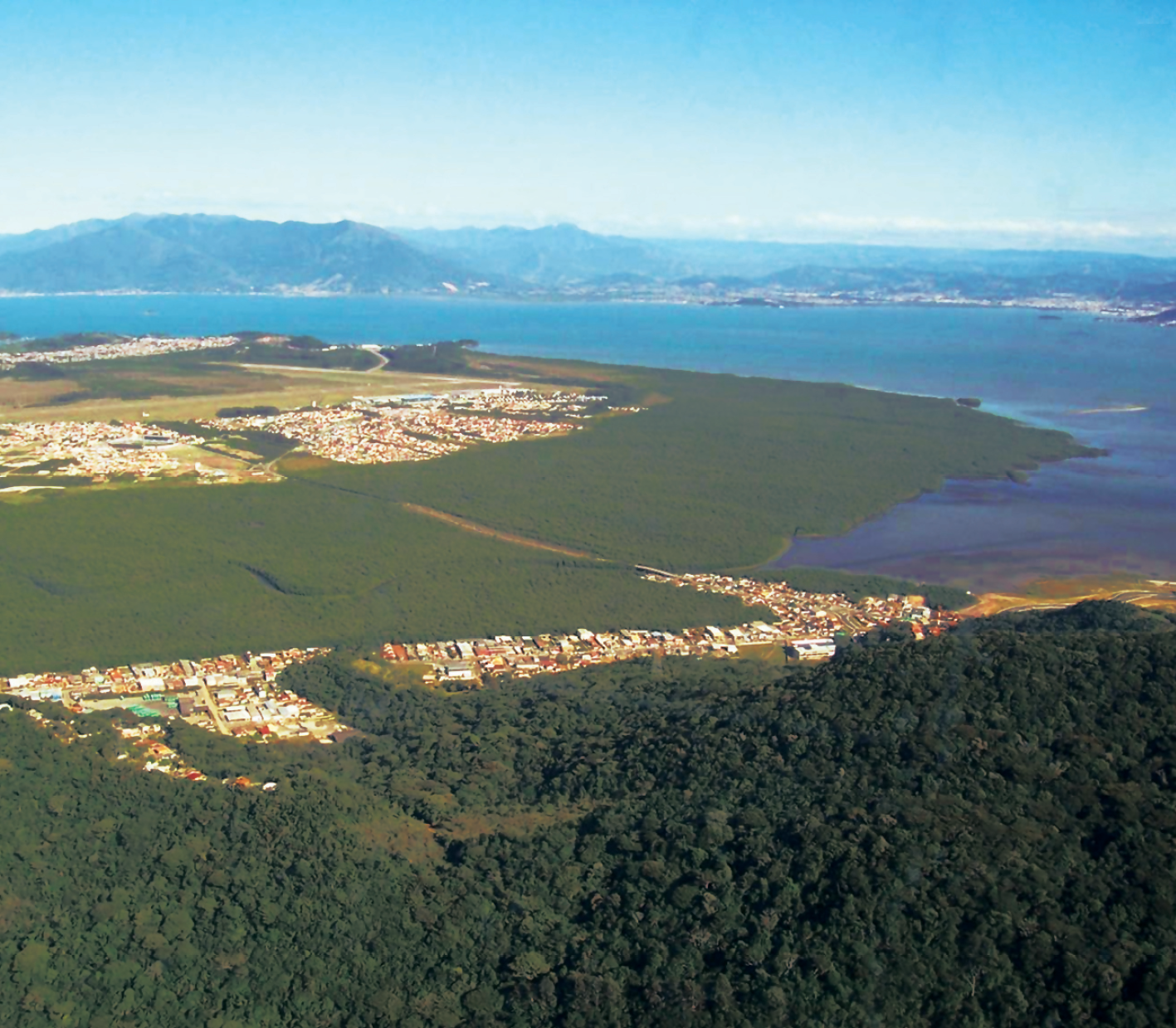 Fotografia. Vista do alto mostrando uma parte da ilha de Santa Catarina, com muita vegetação e algumas construções esparsas. Na parte de cima da fotografia aparece o mar (a Baia Sul). Ao fundo, na parte de cima, vista do continente com alguns morros em destaque.