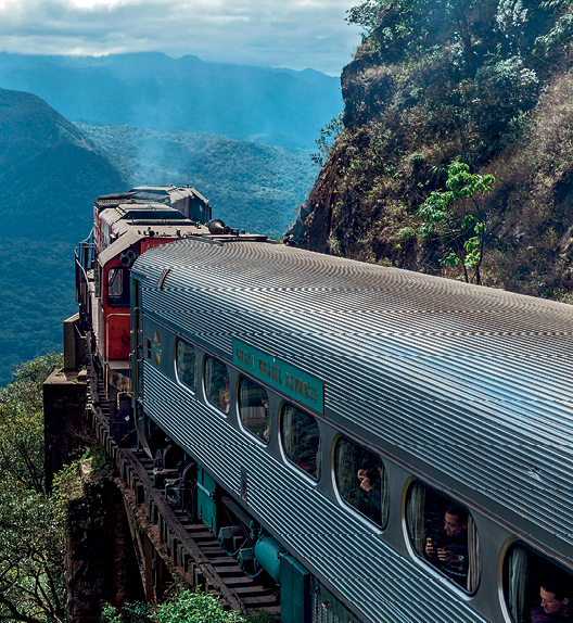 Fotografia. Vista de um trem com vagões de passageiros se deslocando por uma região montanhosa, com terreno acidentando com declividades acentuadas. À esquerda do trem, um grande abismo.
