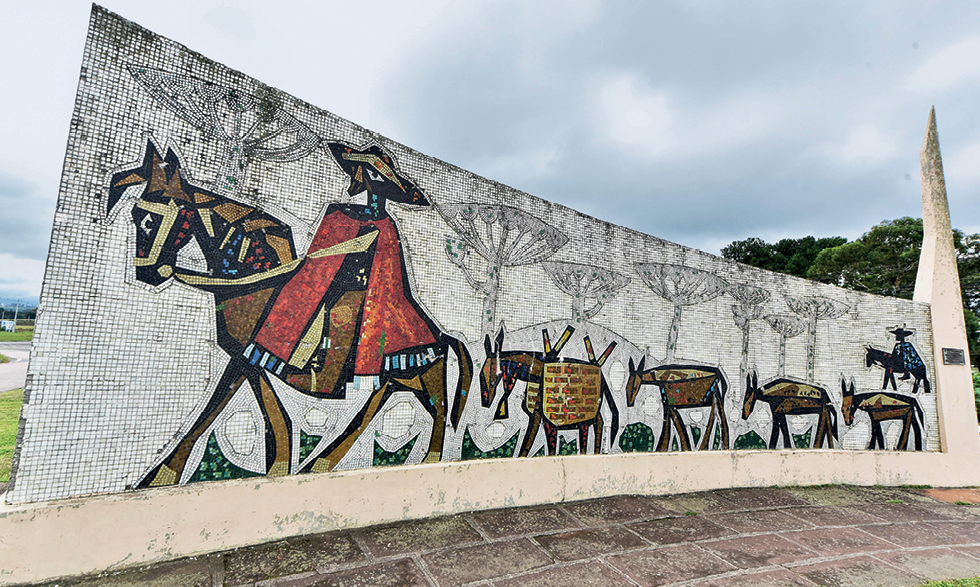 Mural. Mosaico feito em um muro curvado representando a passagem de tropeiros e sua tropa de mulas. À esquerda, um homem de chapéu, usando um manto vermelho e montado em um cavalo. Atrás dele, uma fileira de quatro animais representando as mulas usadas para o transporte de cargas. À direita, mais um homem montado em um cavalo, ele está de chapéu e usa um manto azul.