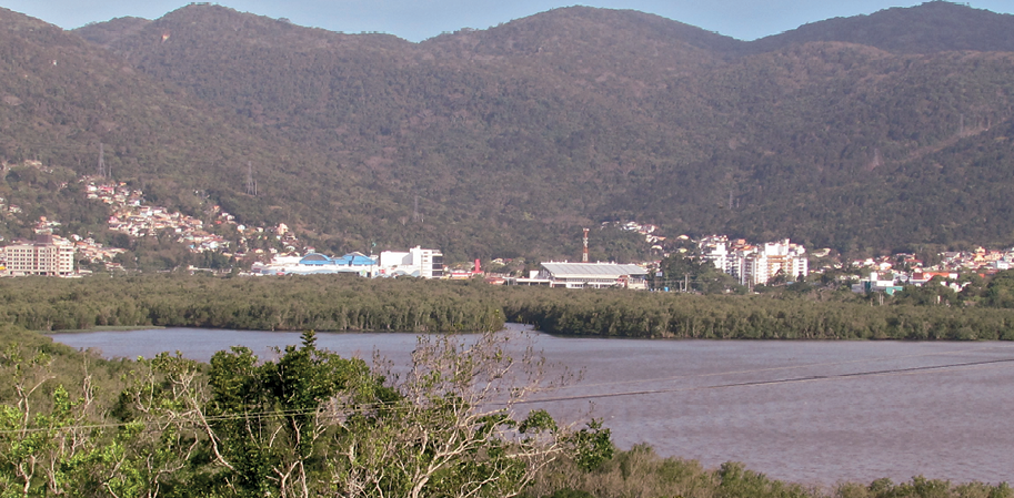 Fotografia. No primeiro plano, vista de uma área de relevo aplainado, com um rio e vegetação nas margens. No segundo plano, vista de construções e morros com vegetação; há construções também na parte de baixo de um dos morros, à esquerda, em uma área de declividade acentuada.