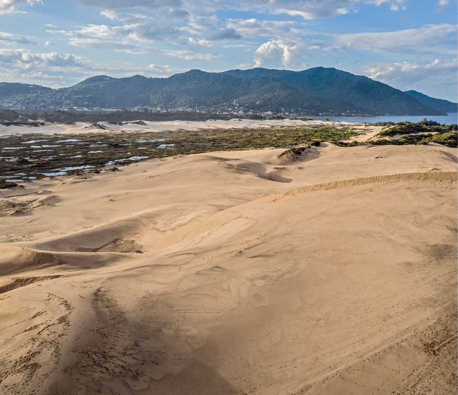 Fotografia. No primeiro plano, vista de uma área extensa formada por dunas entremeadas por uma faixa de restinga; no segundo plano, à direita, uma vista do mar; no terceiro plano, vista de uma área de morros.
