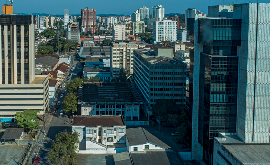 Fotografia. Vista do alto de algumas quadras de uma cidade, com ruas se cruzando e uma concentração de edifícios altos, de vários andares.