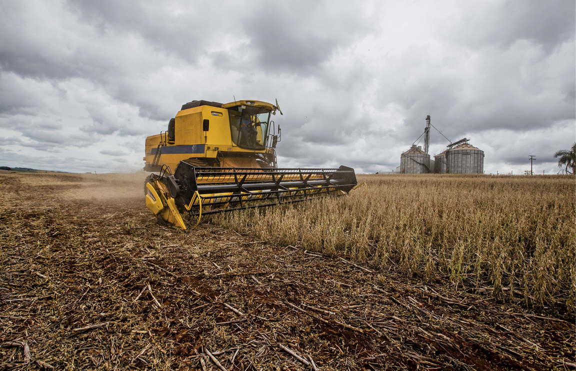 Fotografia. No primeiro plano, vista de uma máquina agrícola em uma área de plantio. No segundo plano, vista de um silo, uma estrutura de metal usada para armazenagem de grãos.