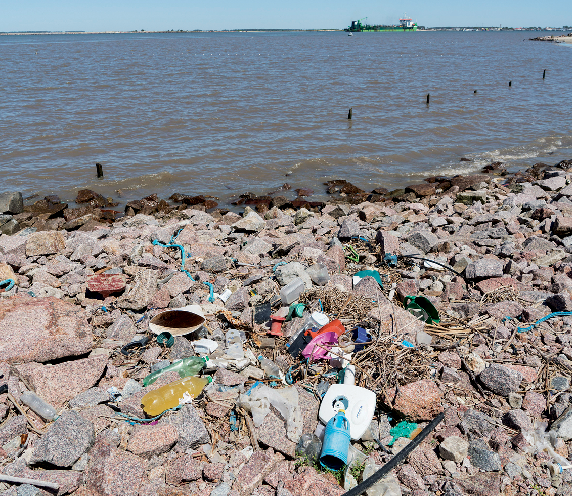 Fotografia. No primeiro plano, vista da margem de um lago, com lixo acumulado próximo ao corpo de água. No segundo plano, o lago e uma embarcação ao fundo.