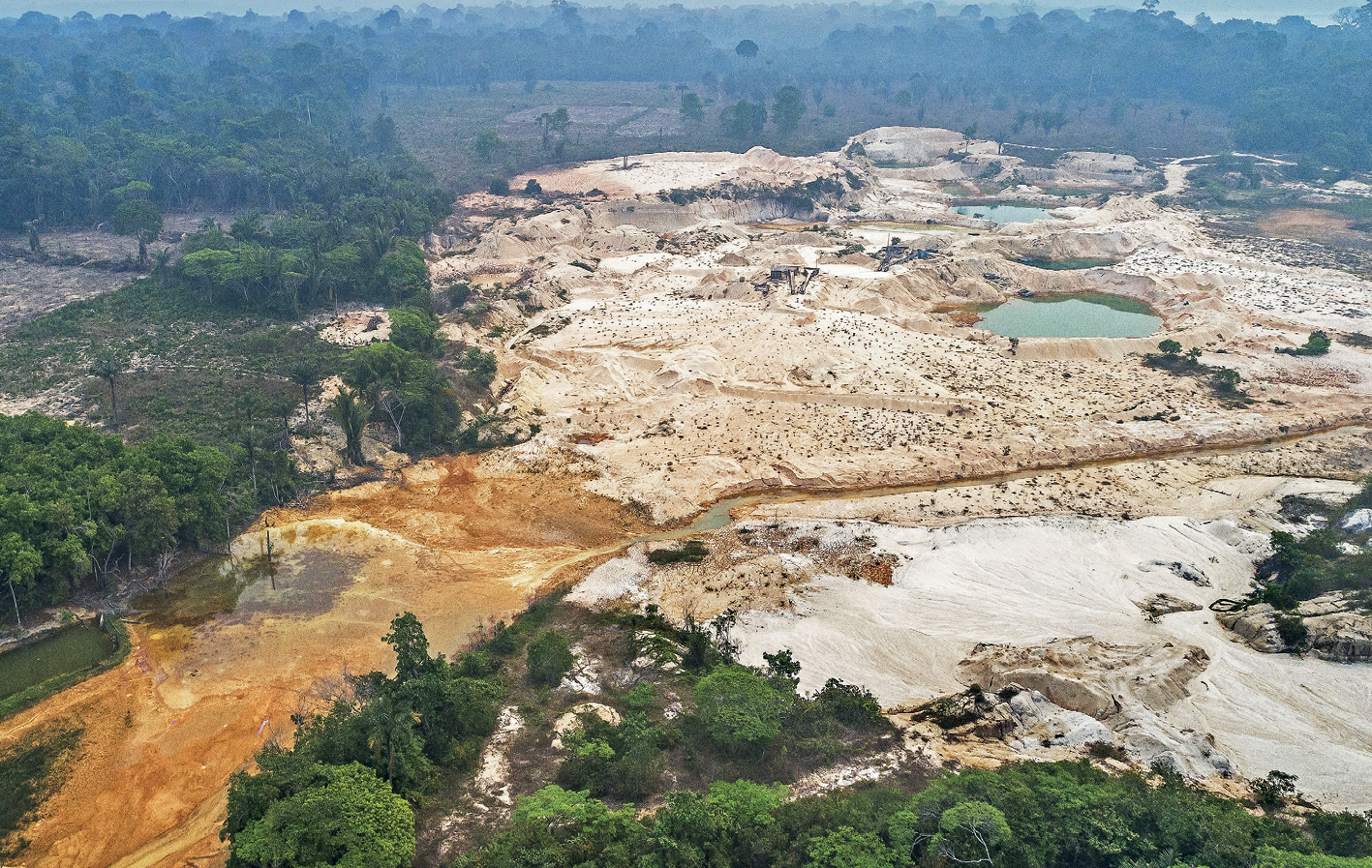 Fotografia. Na parte central e inferior esquerda, vista de uma extensa área acidentada e com solo exposto, em tons de bege e marrom, e com alguns buracos de tamanho mediano preenchidos por água. Ao redor dessa área, há árvores que, em alguns locais estão próximas e, em outros, mais esparsadas.