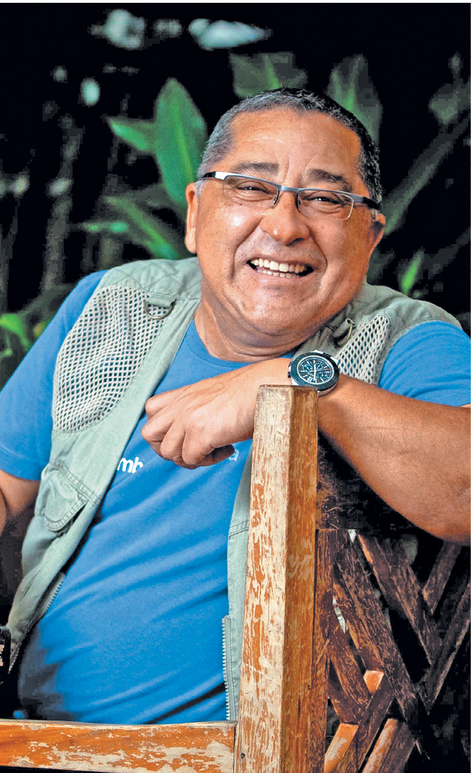 Fotografia de um homem sentado em uma cadeira de madeira. Ele possui cabelos curtos e grisalhos e utiliza óculos de grau, camiseta azul e colete verde. Ele sorri para foto.