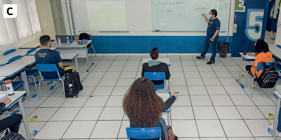 Fotografia C. Interior de uma sala de aula com alguns alunos sentados e enfileirados em cadeiras com mesas. O professor está explicando algo na lousa.