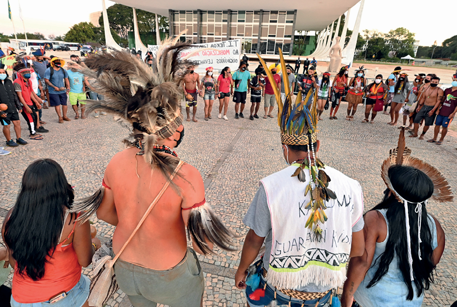 Fotografia. Em uma praça,  há uma roda de pessoas indígenas, a maioria está de mãos dadas. Alguns usam cocar na cabeça, objeto feito de penas. Todos usam máscara. Ao fundo uma grande construção com pilares e grandes paredes de vidros.