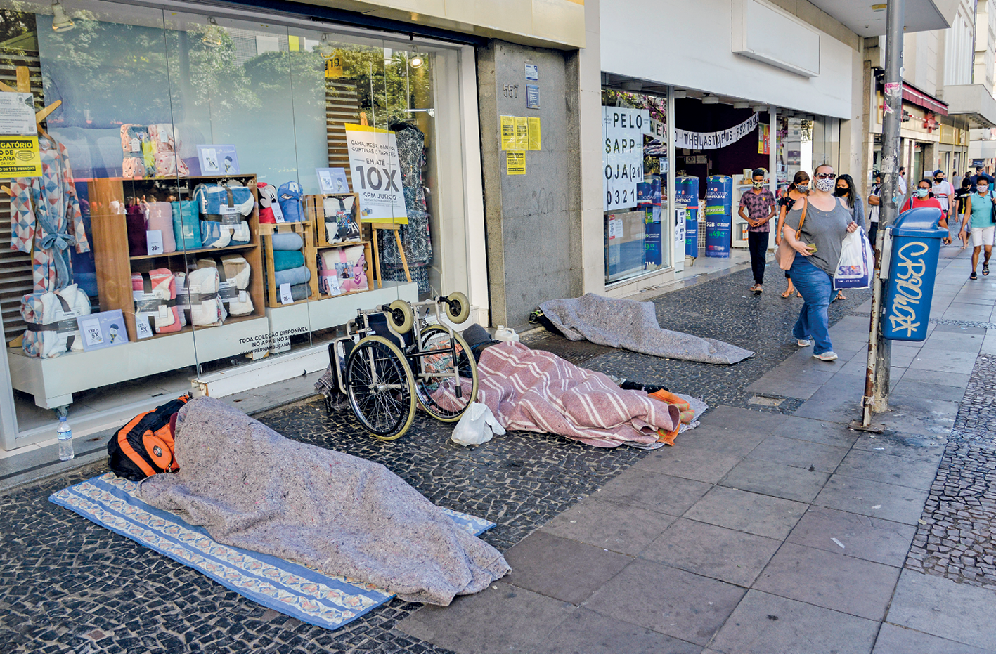 Fotografia. Vista de uma calçada com três pessoas deitadas. Elas estão encobertas por cobertores. Acima de onde elas estão há diversas lojas e, ao fundo, pessoas andando na mesma calçada.