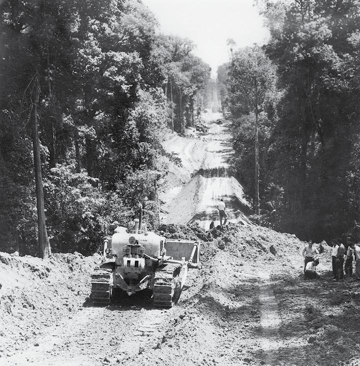 Fotografia em preto e branco. Vista de uma estrada de terra, cercada por árvores altas. Na estrada há um trator e um pequeno monte de terra.