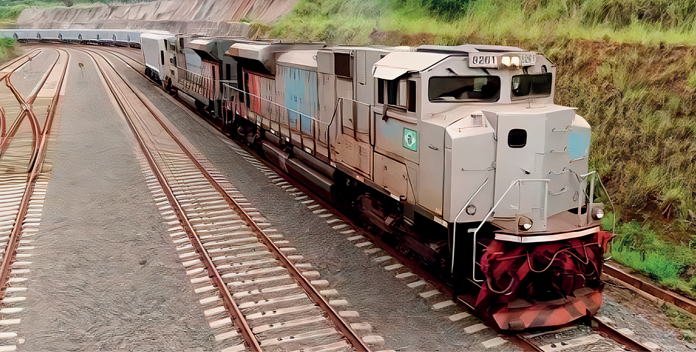 Fotografia. Vista de um trem com alguns vagões em uma linha férrea. Na parte direita da imagem, terreno elevado com vegetação rasteira.