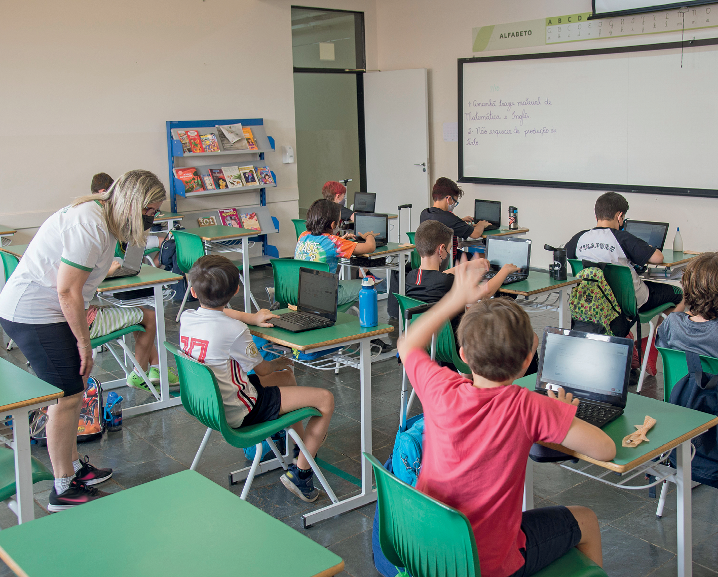 Fotografia. Vista de uma sala de aula com alunos sentados em carteiras verdes enfileiradas. Cada aluno está com um notebook apoiado sobre a mesa. Uma mulher conversa com um dos alunos. Ao fundo, prateleira com livros, uma lousa branca e uma porta.