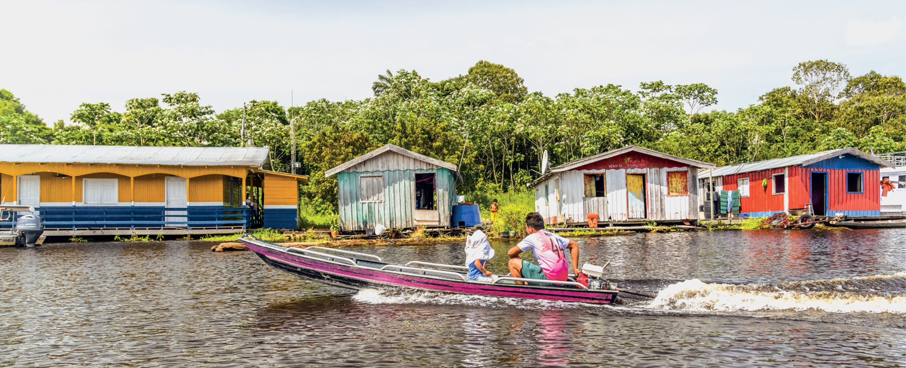 Fotografia. Vista de frente de um rio. No primeiro plano, há um barco pequeno nas cores branco, azul e rosa passando pelo rio com duas pessoas. No segundo plano, casas de madeira e coloridas na beira do rio. Atrás das casas, muitas árvores.