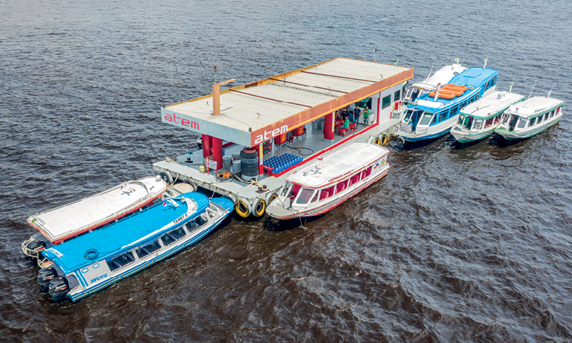 Fotografia. Vista de cima e de lado de um rio com seis embarcações brancas e azuis pequenas. Elas estão paradas. Entre elas há uma estação flutuante.