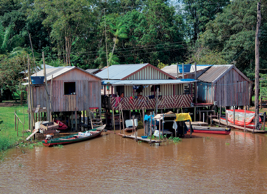 Fotografia. No primeiro plano, rio com coloração marrom e três casas de madeira erguidas sobre estacas. Há pequenos barcos na frente das casas. Ao fundo, muitas árvores.