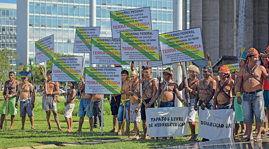 Fotografia. Vista de frente e aproximada de um grupo de indígenas em pé segurando faixas e cartazes. Eles estão em uma área aberta e gramada. Ao fundo, prédios com janelas de vidro.