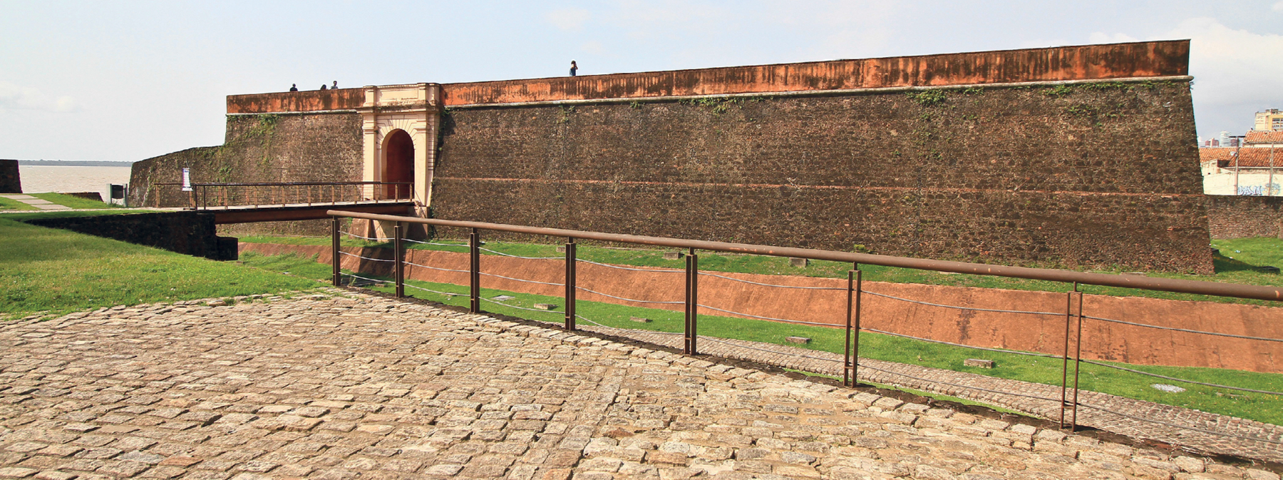Fotografia. Vista de frente da parede de pedras de um forte. O forte tem uma porta de entrada que pode ser acessada por meio de uma ponte de madeira.